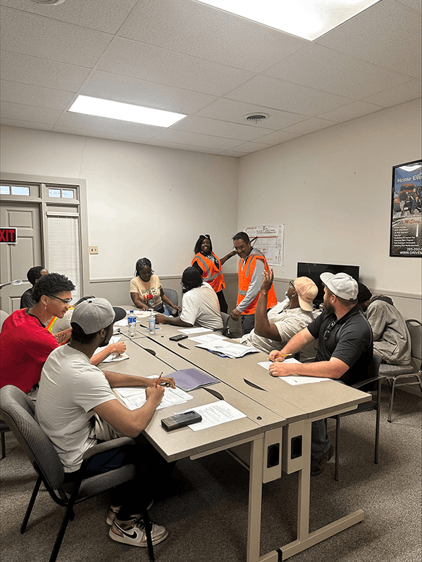 TransTech students sitting at a table with papers. Instructor pointing to poster on the wall.