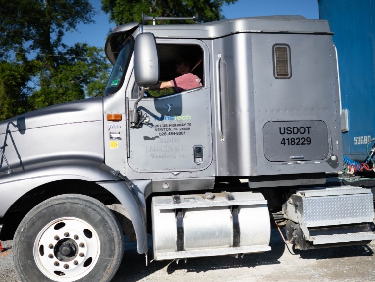 Silver semi truck cab in training yard. Driver sitting in passenger seat with arm out the window.