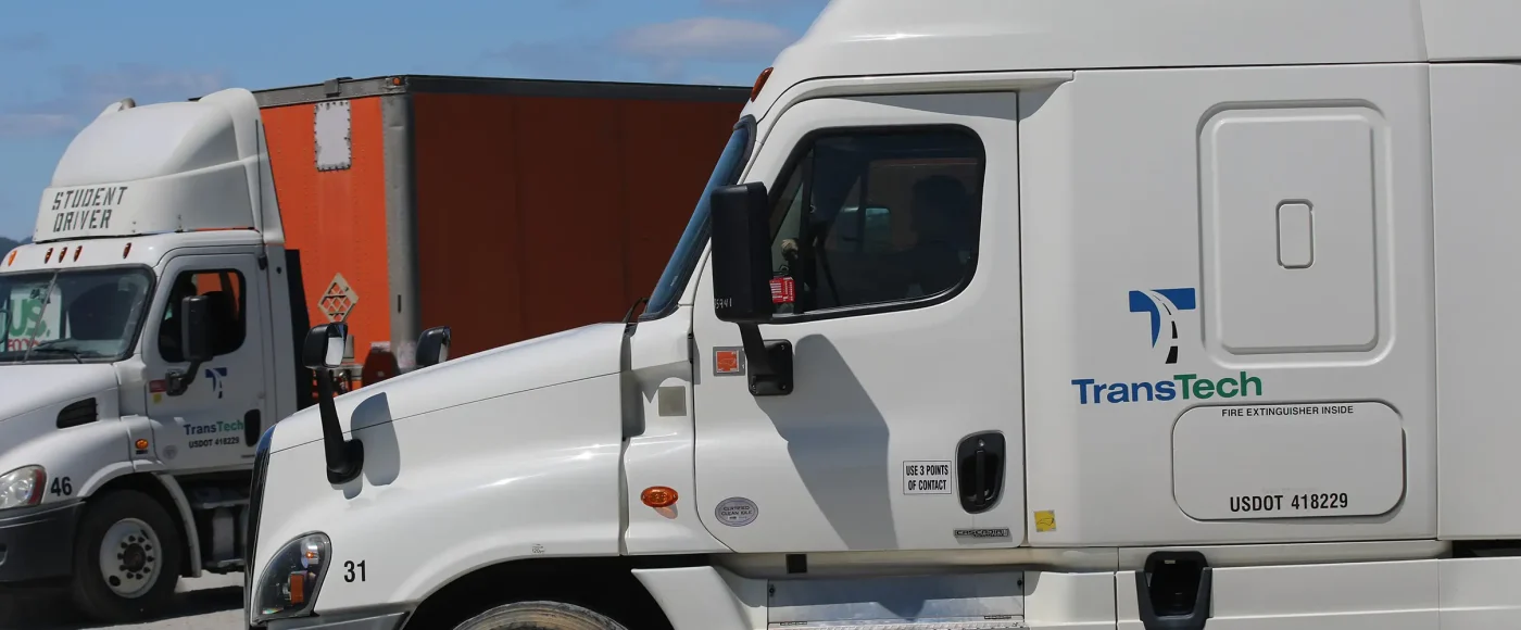 Two TransTech student driver semi-trucks lined up at a CDL training yard, with a student visible through the cab window of the foreground truck