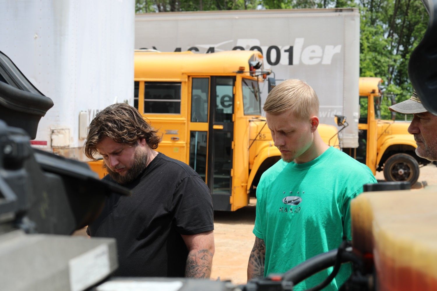 Two CDL students closely observe an under-hood engine inspection during pre-trip training at a truck driving school yard with school buses in the background