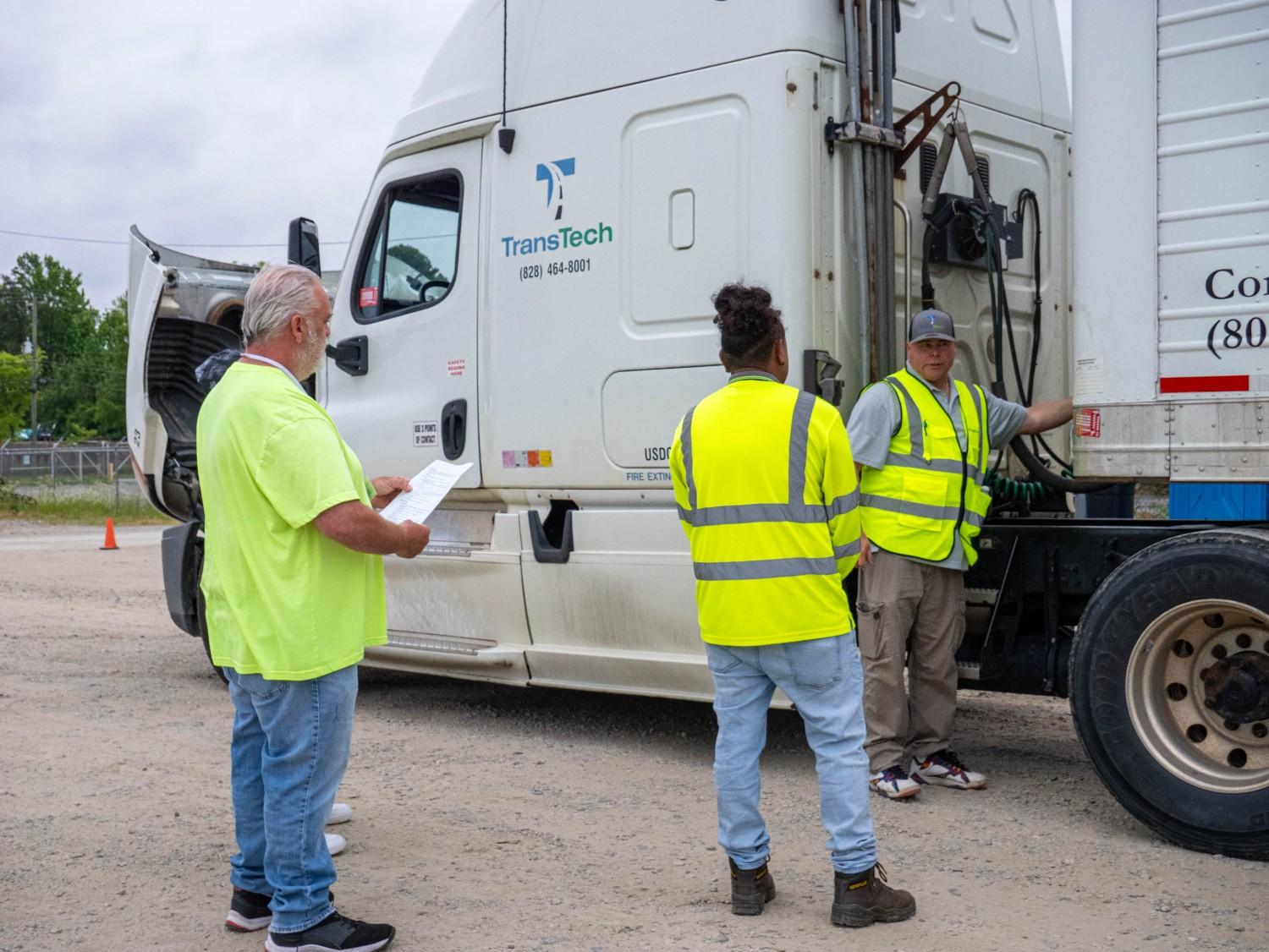 Three TransTech CDL students in safety vests conduct a pre-trip inspection walkthrough on a semi-truck, with one holding a checklist at the training yard