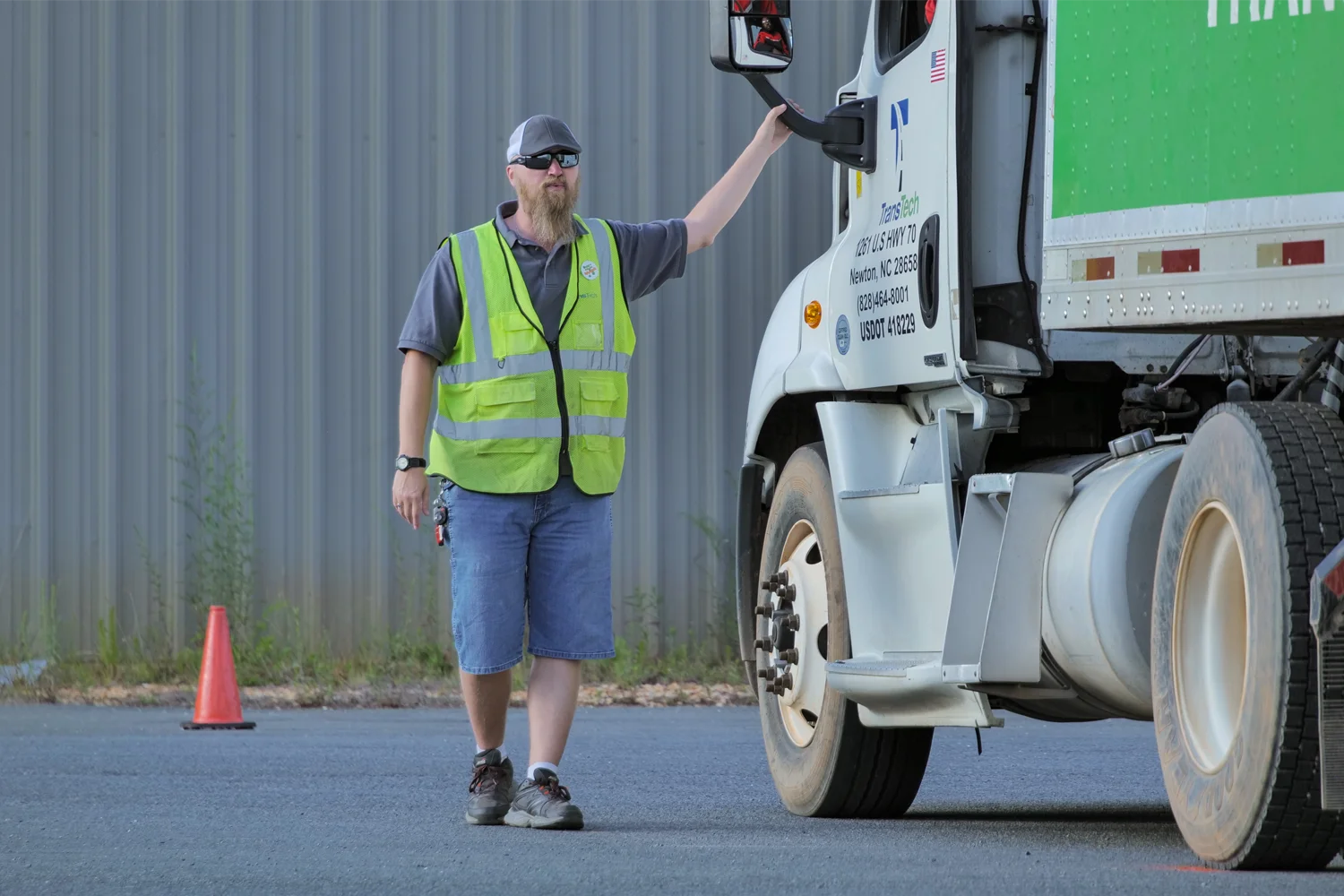 A CDL instructor in a safety vest and sunglasses standing beside a TransTech semi-truck on a training lot, resting a hand on the cab.