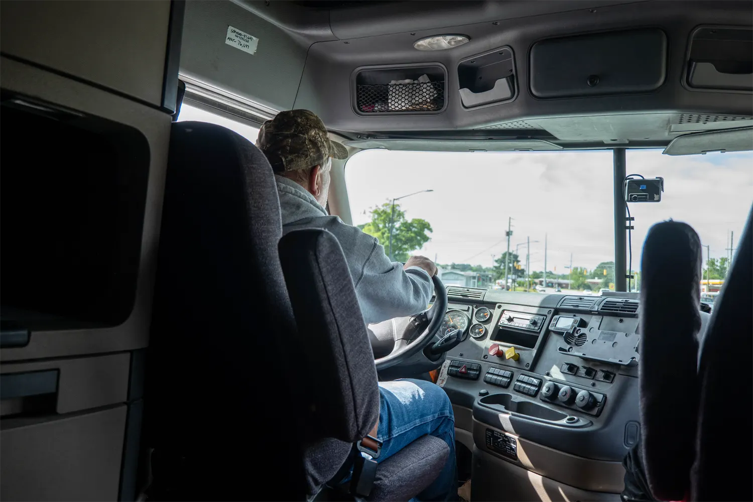 View from inside a semi truck cab, seen from behind the driver's seat, as a driver in a camouflage cap steers through a town intersection with the dashboard and a passenger seat in view.