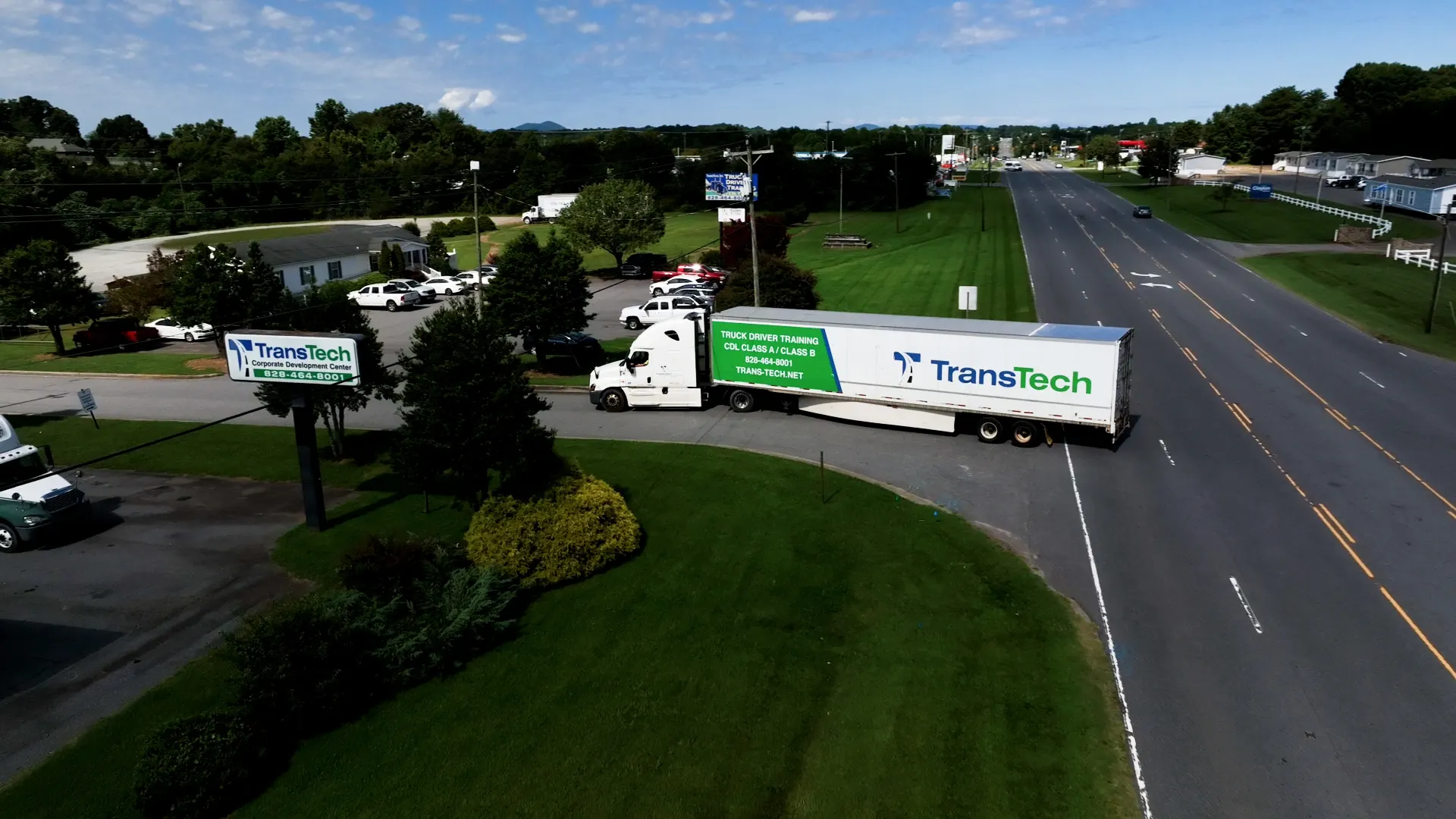 Aerial view of a white TransTech semi truck pulling out of the TransTech Corporate Development Center onto a multi-lane road, with green lawns, trees, and distant mountains in the background.